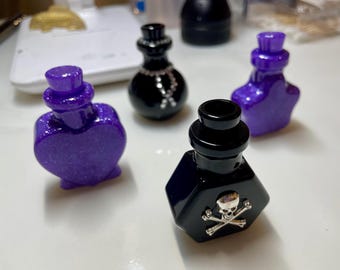 May include: An assortment of decorative potion bottles in various shapes and colors. The bottles are black and purple, with glitter accents. One bottle features a skull and crossbones design, while another has a heart shape. The bottles are likely for decorative purposes.