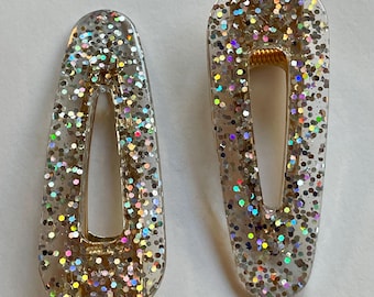 May include: Two silver glitter hair clips with a gold metal spring. The clips are shaped like an oval with a rounded top and bottom.