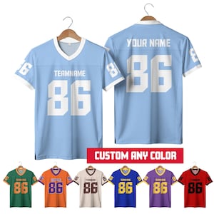 Pode incluir: Camisola de futebol azul claro com detalhes brancos, o número 86 e o texto "TEAMNAME" na frente. A parte de trás da camisola tem o número 86 e o texto "YOUR NAME". Camisolas adicionais em verde, laranja, bege, azul, roxo e vermelho são exibidas.