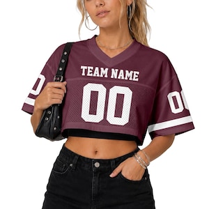 Pode incluir: Uma camisola curta bordô com decote em V, texto branco TEAM NAME e o número 00 na frente. A peça de manga curta apresenta uma textura de malha no peito e riscas brancas nas mangas, combinada com uma saia de ganga preta.