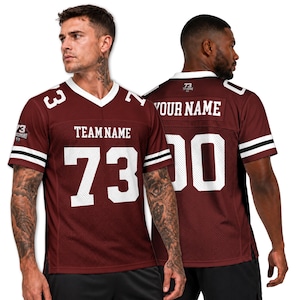 Puede incluir: Una camiseta de fútbol americano color granate con cuello en V blanco y rayas blancas en las mangas. El frente presenta el texto TEAM NAME y el número 73 en blanco. La espalda muestra el texto YOUR NAME y el número 00 en blanco, con un logo 73 en el cuello.