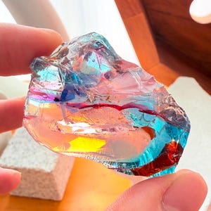 Puede incluir: Un trozo de vidrio bruto e irregular con reflejos iridiscentes, que presenta un espectro de colores vibrantes que incluye azul intenso, amarillo brillante y naranja cálido. La superficie es translúcida con bordes afilados y un acabado reflectante.