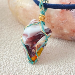 May include: A pendant featuring a multi-colored glass stone with swirls of white, deep red, and yellow. The stone is wrapped in a combination of thin gold-colored wire and teal-colored cord, which also forms the bail attached to a dark blue cord necklace.