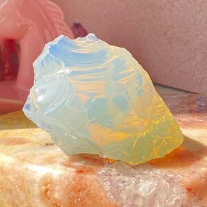 Puede incluir: Una piedra de opalita cruda y translúcida con una textura suave similar al vidrio y bordes irregulares y dentados. La piedra muestra una coloración azul pálido y blanco lechoso con sutiles reflejos de luz amarillos y naranjas.