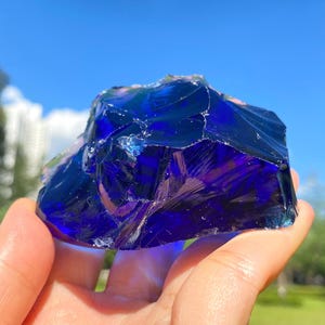 Può includere: Un grande pezzo irregolare di vetro blu cobalto profondo con un aspetto traslucido e cristallino. La superficie presenta bordi affilati e frastagliati e lisce fratture concoidali che catturano la luce, creando una tonalità blu vibrante.
