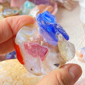 Può includere: Una pietra decorativa circolare composta da frammenti di vetro fuso in tonalità vivaci di blu cobalto, rosa traslucido, arancione brillante e bianco latte. La superficie presenta un mix di texture lisce e frastagliate con inclusioni stratificate.