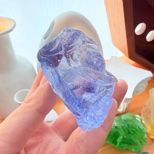 Puede incluir: Una roca de vidrio translúcido de color violeta claro con una forma irregular y dentada, que presenta burbujas de aire internas visibles. La pieza tiene una superficie lisa y reflectante con bordes afilados y angulosos en todo el material.