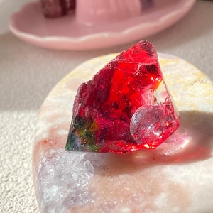 Puede incluir: Un trozo de vidrio rojo translúcido y dentado con fracturas internas y sutiles toques de tonos verdes y amarillos. El fragmento de vidrio descansa sobre una piedra lisa de forma ovalada con una textura moteada en blanco, rosa y beige.