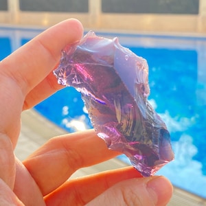 Puede incluir: Un trozo de vidrio morado translúcido e irregular con una superficie lisa y reflectante, además de bordes afilados y dentados. El material capta la luz, mostrando tonos violetas profundos y secciones claras que permiten el paso de la luz.
