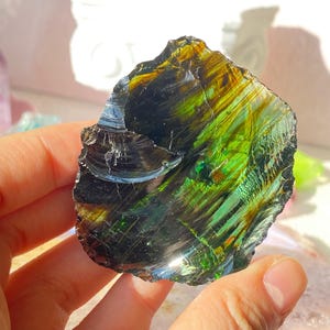 Puede incluir: Una losa irregular de vidrio obsidiana pulido que muestra un brillo iridiscente vibrante con vetas de verde bosque profundo, amarillo dorado y negro carbón. La superficie es lisa y reflectante, mostrando fracturas concoideas naturales.