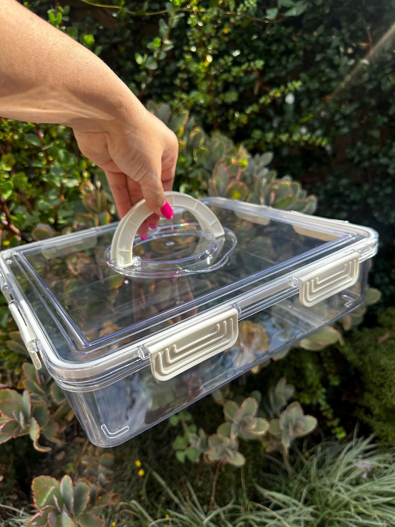 Puede incluir: Un contenedor de almacenamiento de pl&aacute;stico transparente con una manija blanca y cierres dorados. El contenedor tiene una tapa transparente.