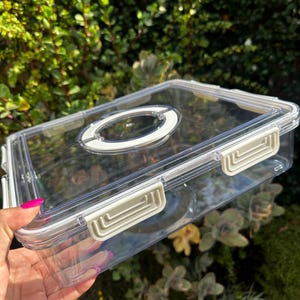 Puede incluir: Un recipiente de almacenamiento de pl&aacute;stico transparente con una tapa blanca y un cierre blanco en el lateral. El recipiente tiene una manija redonda transparente en la parte superior.