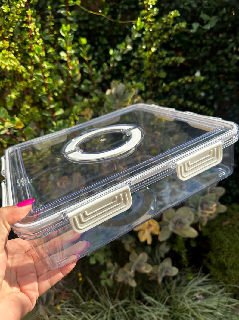 Puede incluir: Un recipiente de almacenamiento de pl&aacute;stico transparente con una tapa blanca y un cierre blanco en el lateral. El recipiente tiene una manija redonda transparente en la parte superior.