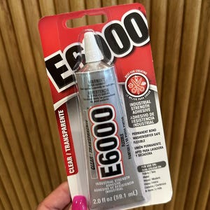 May include: A tube of E6000 industrial strength adhesive, clear and flexible, on a red and white card. The tube contains 59.1 ml of adhesive. The packaging includes warnings and instructions for use on various materials.