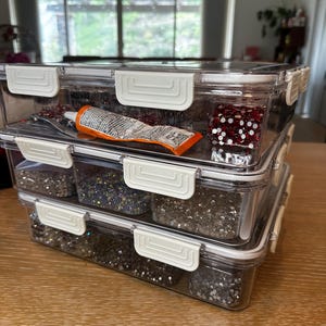 Puede incluir: Tres contenedores de pl&aacute;stico transparente apilados uno encima del otro. El contenedor superior tiene un dado rojo y blanco y un tubo de pegamento. El contenedor del medio tiene purpurina plateada. El contenedor inferior tiene purpurina plateada.