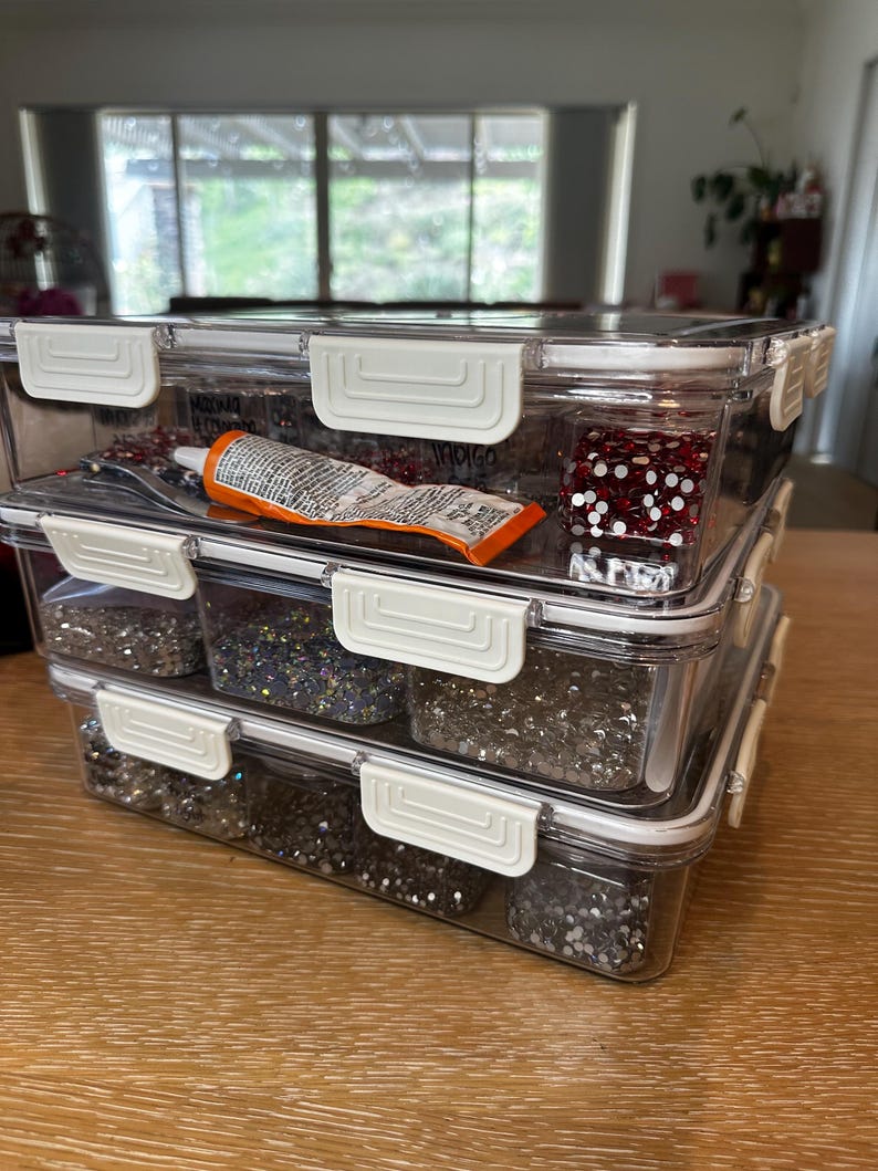 Puede incluir: Tres contenedores de pl&aacute;stico transparente apilados uno encima del otro. El contenedor superior tiene un dado rojo y blanco y un tubo de pegamento. El contenedor del medio tiene purpurina plateada. El contenedor inferior tiene purpurina plateada.