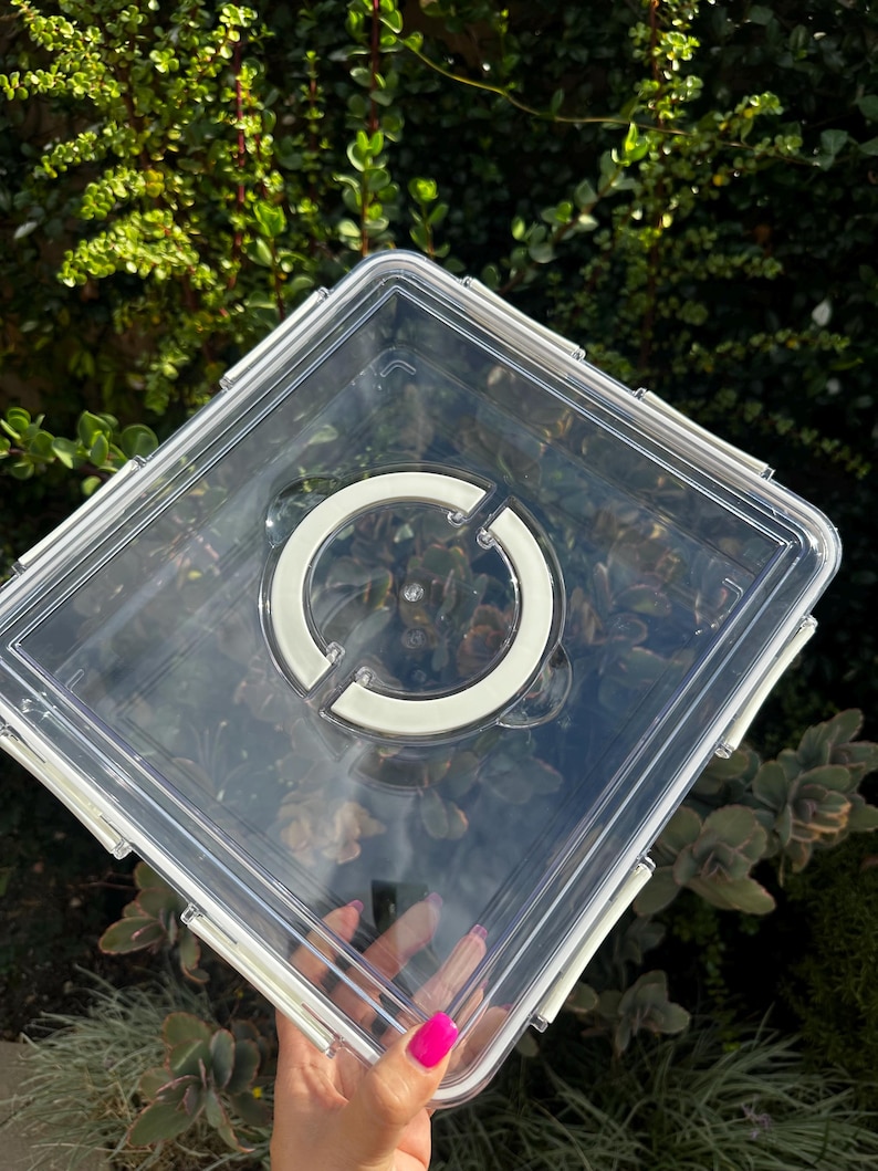 Puede incluir: Un contenedor de almacenamiento de pl&aacute;stico transparente con un cierre blanco y un c&iacute;rculo blanco en la tapa.