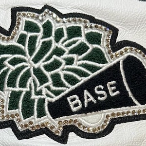 Puede incluir: Parche de chaqueta de cuero blanco con un diseño floral verde y blanco, borde negro y detalles de pedrería. Un megáfono negro con la palabra "BASE" también aparece en el parche.