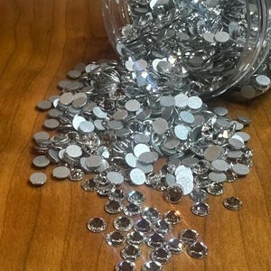 May include: A clear plastic jar tipped over, spilling a large quantity of silver rhinestones onto a wooden surface. The rhinestones are round with a flat back and a faceted, sparkling top. The wooden surface is a warm brown colour.