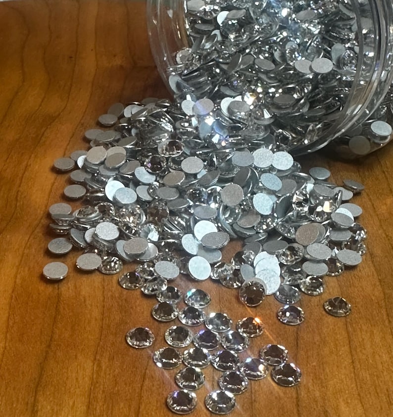 May include: A clear glass jar filled with silver and clear rhinestones. The jar is tipped over, spilling the rhinestones onto a wooden surface.