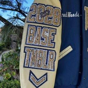 May include: A close-up of a varsity jacket sleeve. The sleeve is tan with navy blue accents and features the text "2028", "BASE", and "TMBLR" in gold and blue rhinestones. The jacket is navy blue.