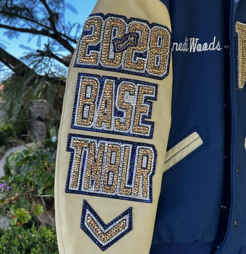 May include: A close-up of a varsity jacket sleeve. The sleeve is tan with navy blue accents and features the text "2028", "BASE", and "TMBLR" in gold and blue rhinestones. The jacket is navy blue.