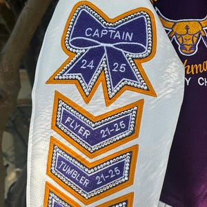 May include: A white and purple varsity jacket with a white leather sleeve. The sleeve has a purple bow with rhinestones and the word "CAPTAIN" on it. There are also four purple patches with rhinestones and the words "FLYER", "TUMBLER", and "BASE" on them.