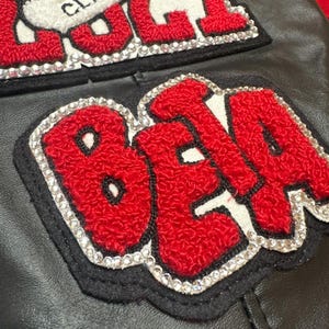 May include: Close-up of a black leather jacket with two embroidered patches. The patches feature red, textured lettering outlined in white, with a border of sparkling rhinestones. The top patch has the word "CLASS" and the bottom patch has the word "BETA".