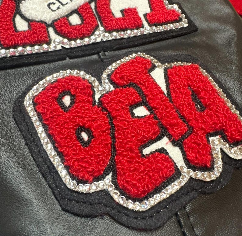 May include: Close-up of a black leather jacket with two embroidered patches. The patches feature red, textured lettering outlined in white, with a border of sparkling rhinestones. The top patch has the word "CLASS" and the bottom patch has the word "BETA".