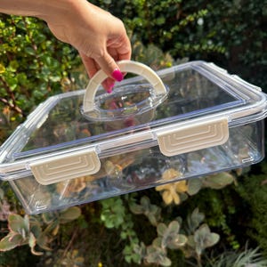 Puede incluir: Un recipiente de almacenamiento de pl&aacute;stico transparente con una tapa blanca y un asa blanca. El recipiente tiene una forma rectangular y est&aacute; hecho de pl&aacute;stico duradero.