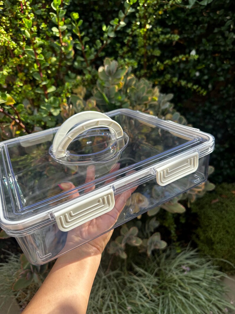 Puede incluir: Un recipiente de almacenamiento de pl&aacute;stico transparente con una tapa blanca y un asa. El recipiente tiene cuatro cierres en los lados.