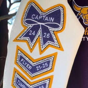 May include: Close-up of white fabric with embroidered patches. The patches include a navy blue bow with the word "CAPTAIN" and the numbers 24 and 25. Other patches say "FLYER" and "TUMBLER" with numbers. All patches have gold borders and rhinestone accents.