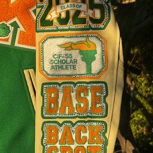 May include: A green and orange varsity jacket with a white leather sleeve. The sleeve has several patches, including a green and orange patch with the text "CLASS OF 2025", a green and orange patch with the text "CIF-SS SCHOLAR ATHLETE", a green and orange patch with the text "BASE", and a green and orange patch with the text "BACK SPOT".