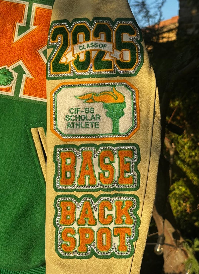 May include: A green and orange varsity jacket with a white leather sleeve. The sleeve has several patches, including a green and orange patch with the text "CLASS OF 2025", a green and orange patch with the text "CIF-SS SCHOLAR ATHLETE", a green and orange patch with the text "BASE", and a green and orange patch with the text "BACK SPOT".