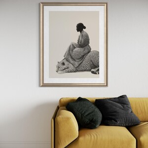 May include: Framed art print featuring a woman seated on a leopard. The artwork is in black and white, with the woman and leopard detailed with a spotted pattern. The frame is gold, and the print is displayed above a yellow sofa with black pillows.