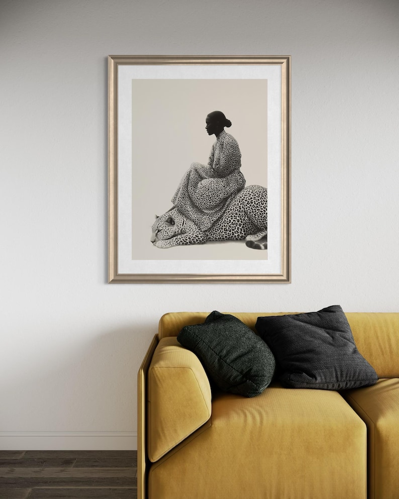 May include: Framed art print featuring a woman seated on a leopard. The artwork is in black and white, with the woman and leopard detailed with a spotted pattern. The frame is gold, and the print is displayed above a yellow sofa with black pillows.