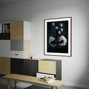May include: A framed art print featuring a dark background with a central figure adorned with white fabric and flowers. The artwork is displayed on a white wall above a wooden bench and modern storage unit.