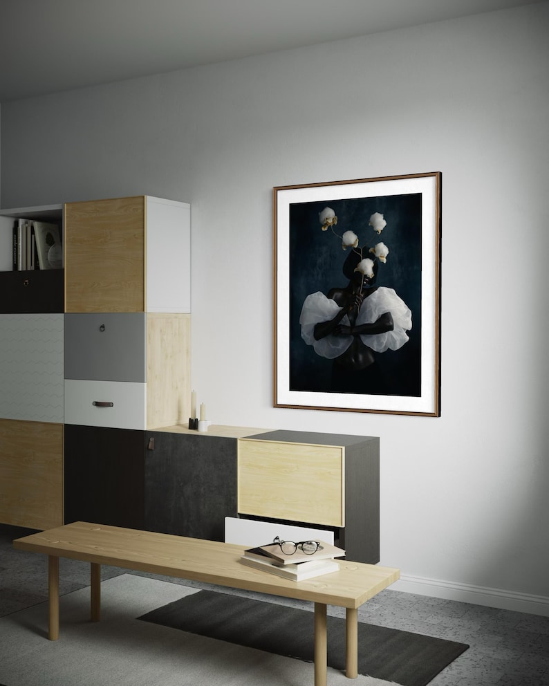 May include: A framed art print featuring a dark background with a central figure adorned with white fabric and flowers. The artwork is displayed on a white wall above a wooden bench and modern storage unit.
