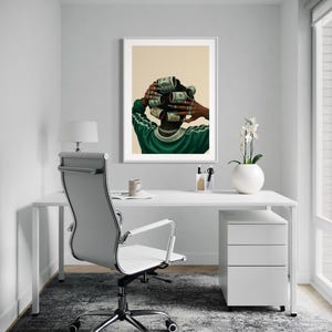 May include: A framed art print depicts a person with dollar bills rolled into curlers. The person wears a green and white track jacket and a gold chain. The artwork hangs above a white desk with a modern office chair and a white filing cabinet.