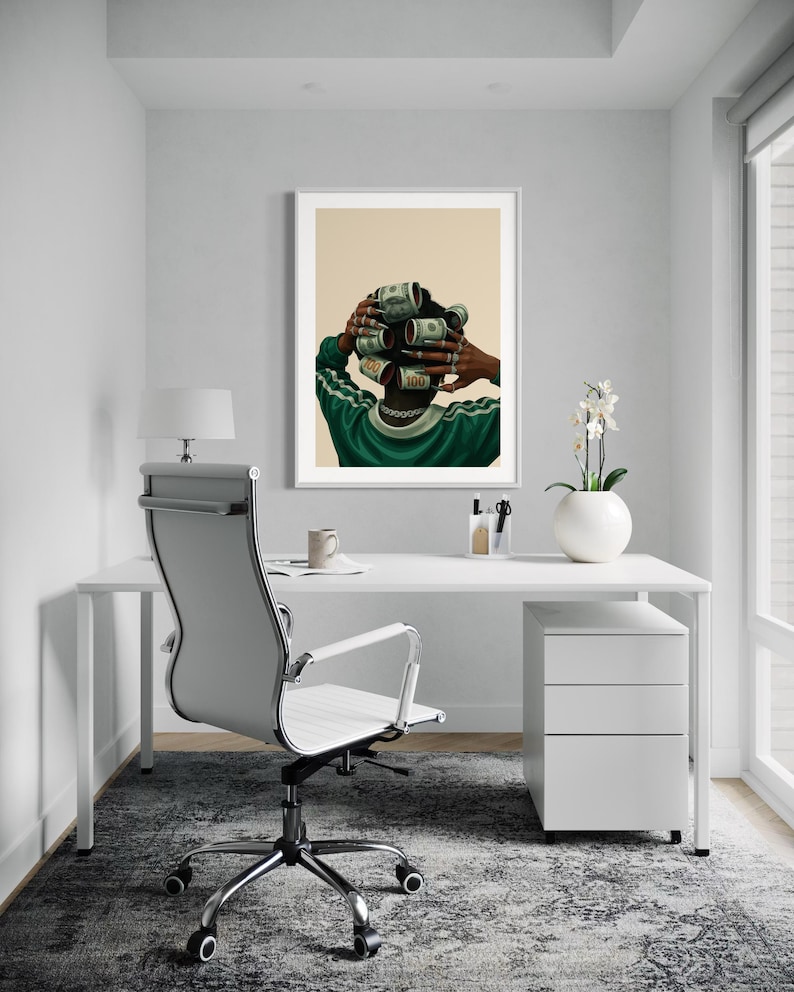 May include: A framed art print depicts a person with dollar bills rolled into curlers. The person wears a green and white track jacket and a gold chain. The artwork hangs above a white desk with a modern office chair and a white filing cabinet.