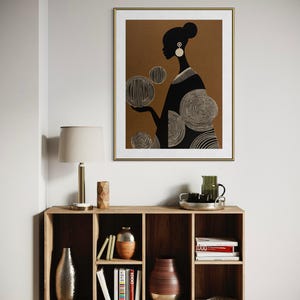 May include: A framed art print featuring a black silhouette of a person with a bun and large earrings, holding stylized circular shapes. The artwork is set against a brown background with a gold frame. The print hangs above a wooden cabinet.