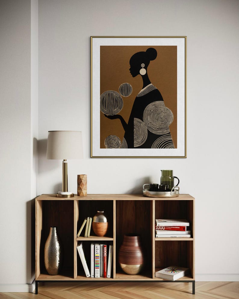 May include: A framed art print featuring a black silhouette of a person with a bun and large earrings, holding stylized circular shapes. The artwork is set against a brown background with a gold frame. The print hangs above a wooden cabinet.