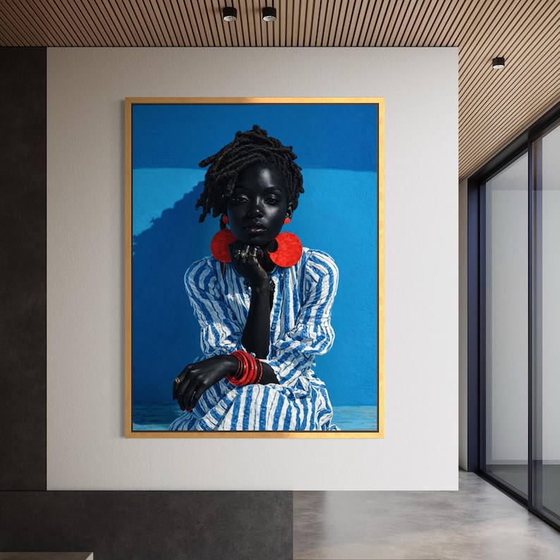 May include: A framed art print showing a portrait of a person with dark skin, wearing a blue and white striped dress, and large red earrings and bracelets. The background is a vibrant blue, and the frame is gold.