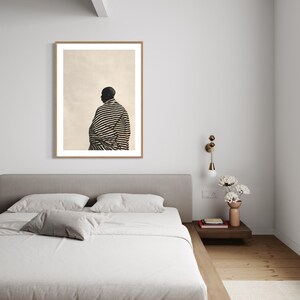 May include: A framed print of a person wearing a black and white striped garment. The artwork is displayed above a bed with a grey headboard and white bedding. A bedside table with a lamp and vase of flowers is next to the bed.