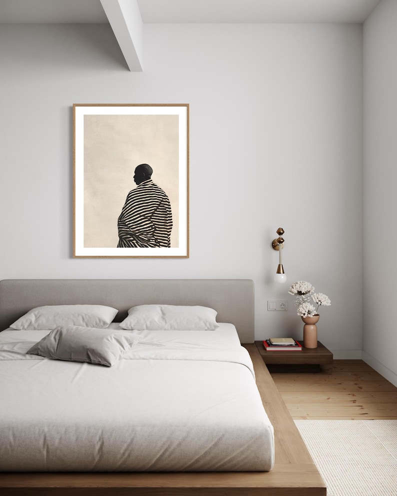 May include: A framed print of a person wearing a black and white striped garment. The artwork is displayed above a bed with a grey headboard and white bedding. A bedside table with a lamp and vase of flowers is next to the bed.