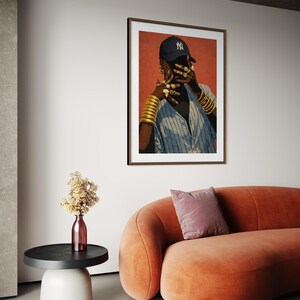 May include: A framed art print featuring a person wearing a navy blue baseball cap with a white logo, gold jewelry, and a blue and white striped shirt. The artwork hangs above an orange curved sofa and a side table.