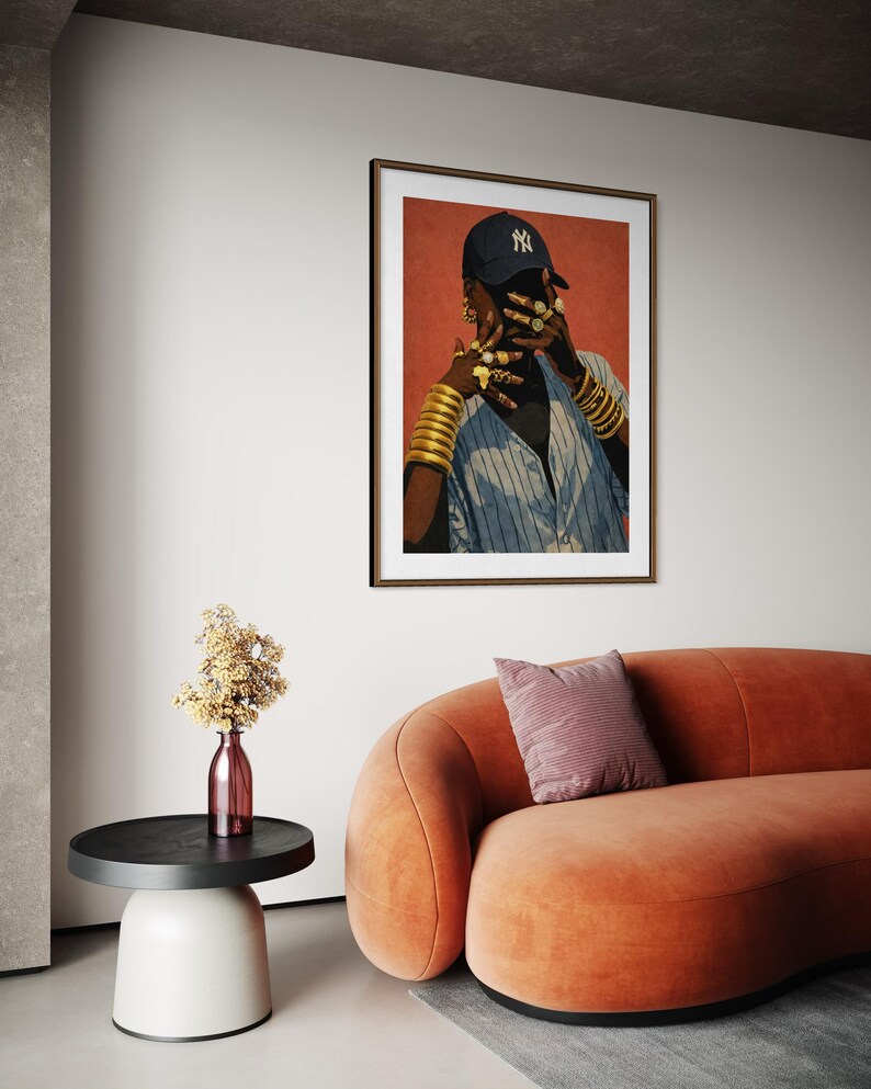 May include: A framed art print featuring a person wearing a navy blue baseball cap with a white logo, gold jewelry, and a blue and white striped shirt. The artwork hangs above an orange curved sofa and a side table.
