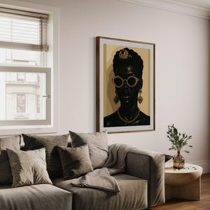 May include: A framed portrait of a woman with dark skin, wearing gold-rimmed sunglasses, a jeweled headpiece, and gold jewelry. The artwork hangs on a cream-colored wall above a gray sofa with decorative pillows and a throw blanket. A small side table with a vase of greenery is next to the sofa.