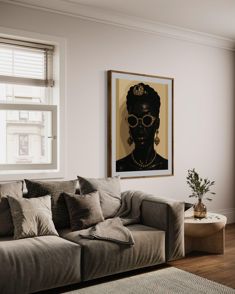 May include: A framed portrait of a woman with dark skin, wearing gold-rimmed sunglasses, a jeweled headpiece, and gold jewelry. The artwork hangs on a cream-colored wall above a gray sofa with decorative pillows and a throw blanket. A small side table with a vase of greenery is next to the sofa.