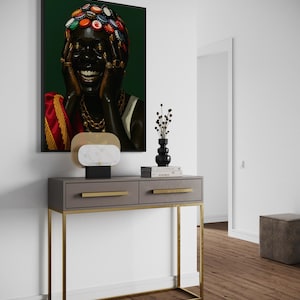 May include: A framed portrait of a person with dark skin, adorned with bottle caps on their head and gold jewelry, laughing. The artwork hangs above a gray console table with gold legs and drawer pulls, complemented by a decorative lamp and vase.